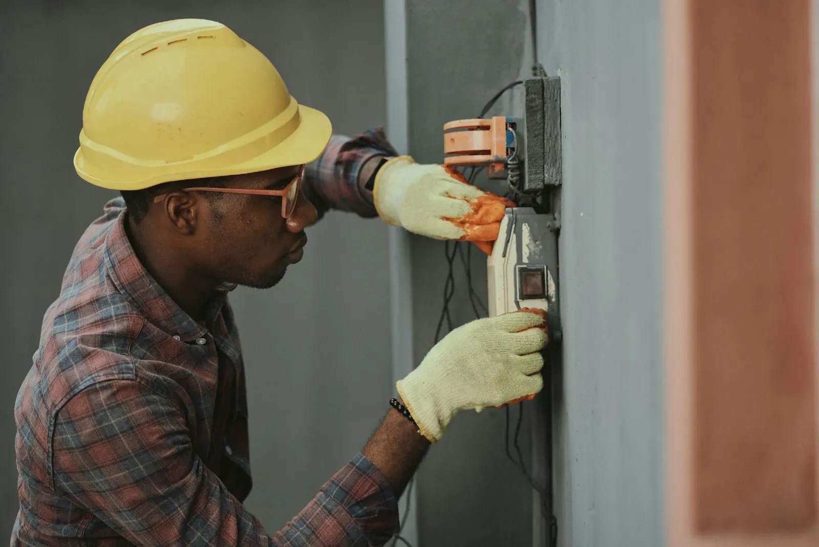 Electrician in a yellow hard hat and safety glasses working on wiring at a wall-mounted meter box